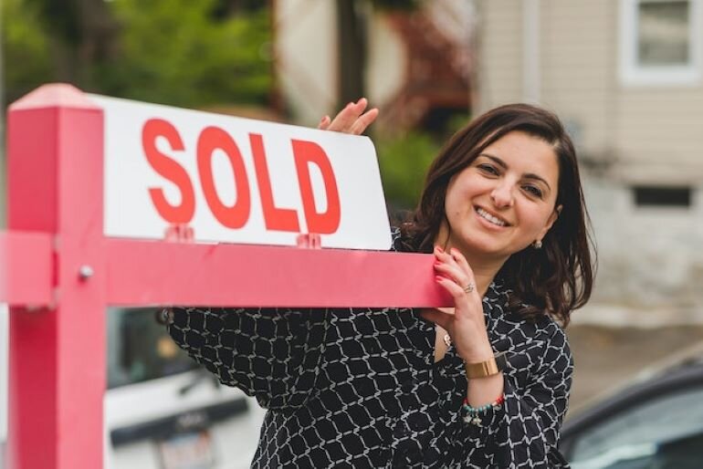 A woman standing next to a house sold sign.