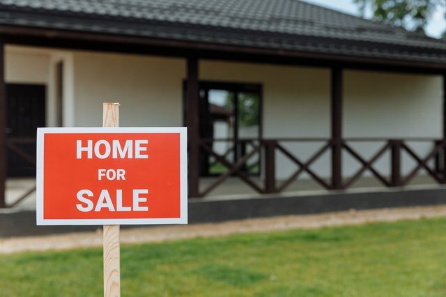 A red and white sign with "home for sale written" on it placed on a lawn in front of a house with a large porch