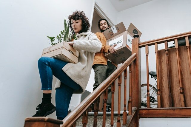 A man and a woman moving out of their apartment and carrying boxes with things down the stairs