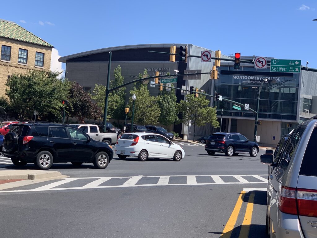 the photo shows an intersection with traffic lights and many vehicles. On the far back of the photo is seen the building of Montgomery College, Takoma Park Maryland