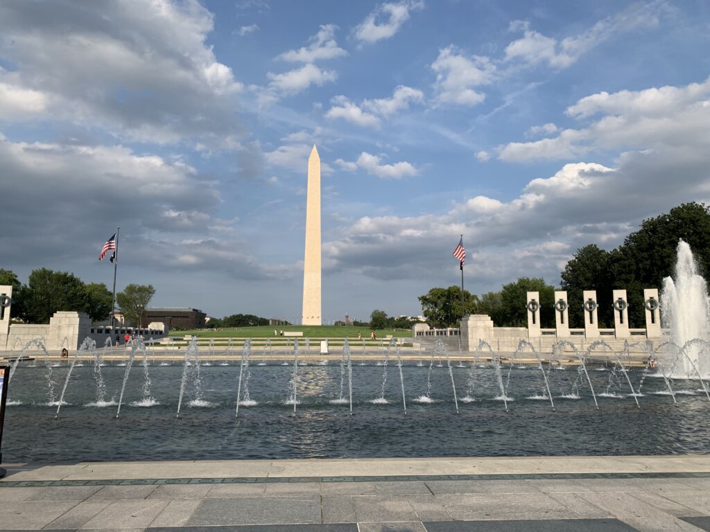 You see a view showing War Word II Memorial with its water fountains and Washington Monument on the far back. Picture also shows blue sky with white and dark clouds.