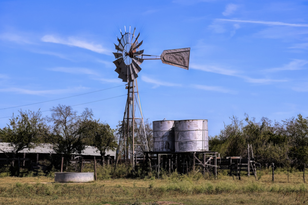 A windmill set in the foreground with 2 water tanks in the background. The landscape surrounding it is lightly forested in a farm-like setting