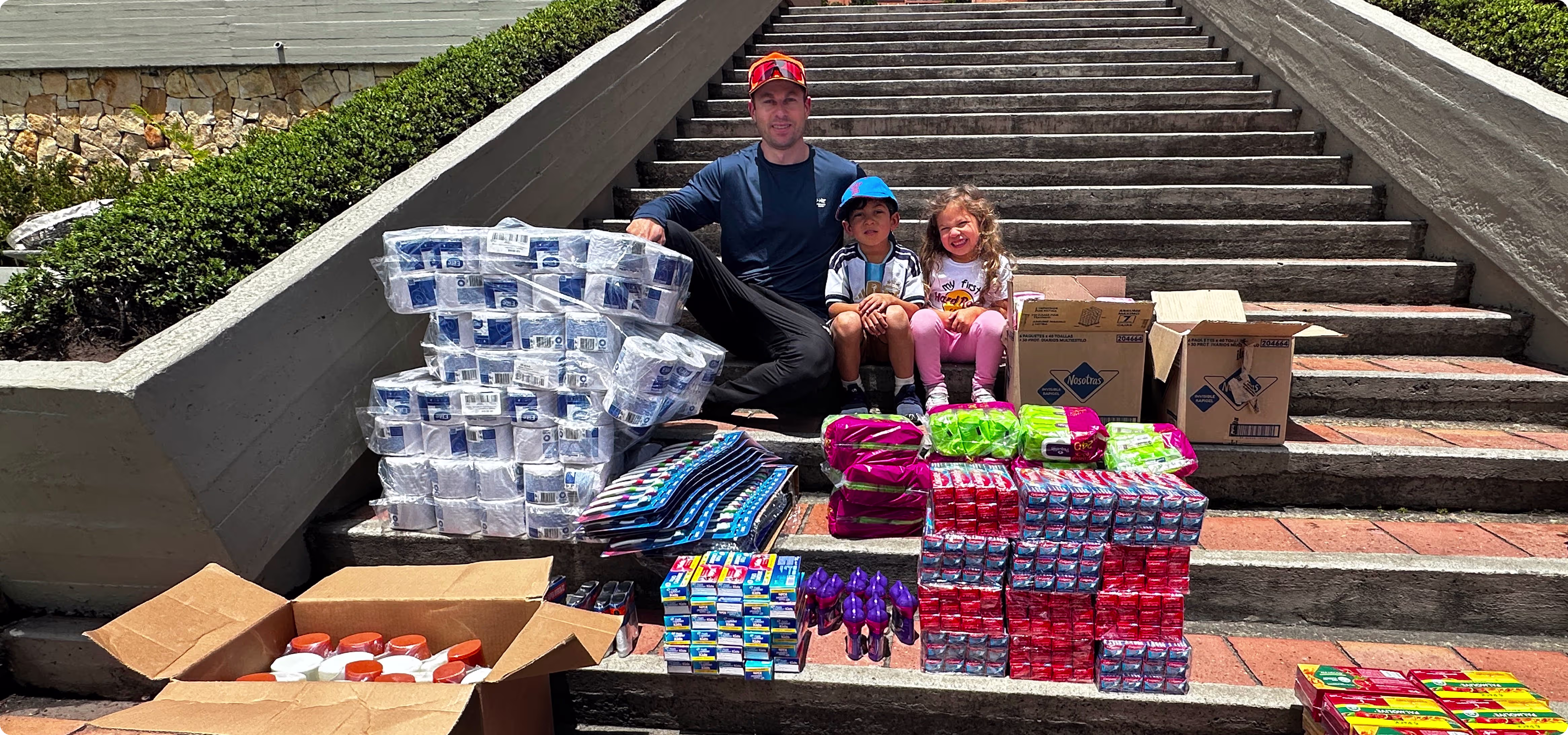 Better Off Home Buyers founder Scott Dalinger and his two children smiling on outdoor stairs at a collection center, surrounded by massive stacks of donated earthquake relief supplies including toilet paper, food, and hygiene products.