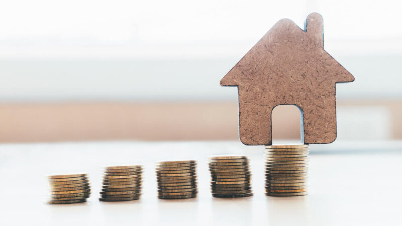 Stack of coins with a house made of cardboard in the background.