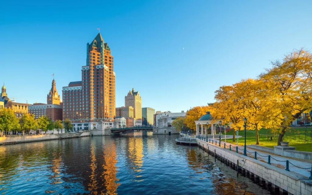 Downtown Milwaukee skyline with tall buildings, autumn trees, and a calm riverfront walkway under a clear blue sky.