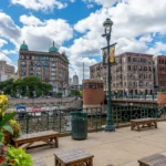 A scenic view of downtown Milwaukee showing historic buildings along the riverwalk with benches, flowers, and a bright blue sky filled with clouds.