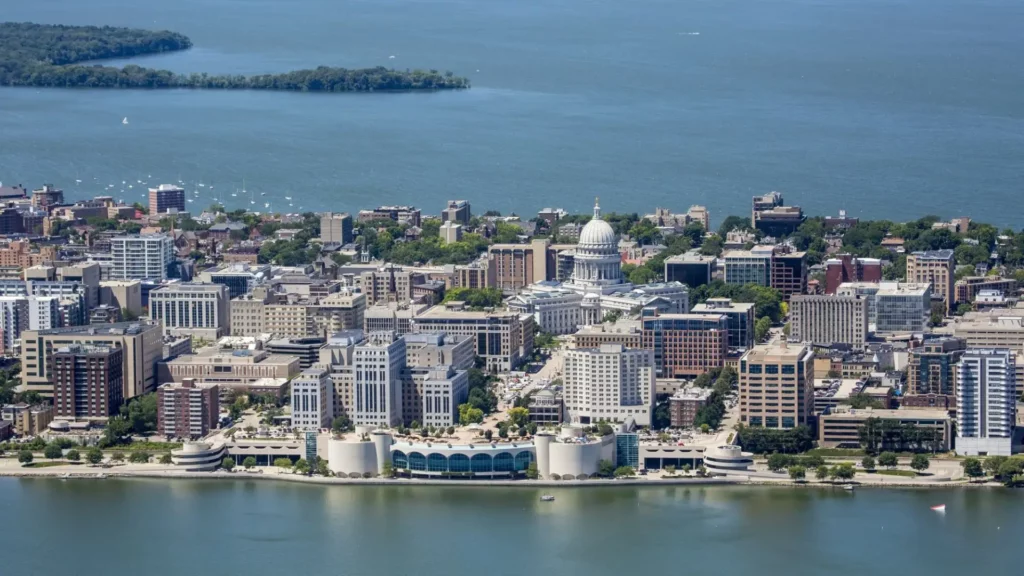 Aerial view of downtown Madison, Wisconsin, featuring the Wisconsin State Capitol surrounded by city buildings and bordered by a large blue lake.